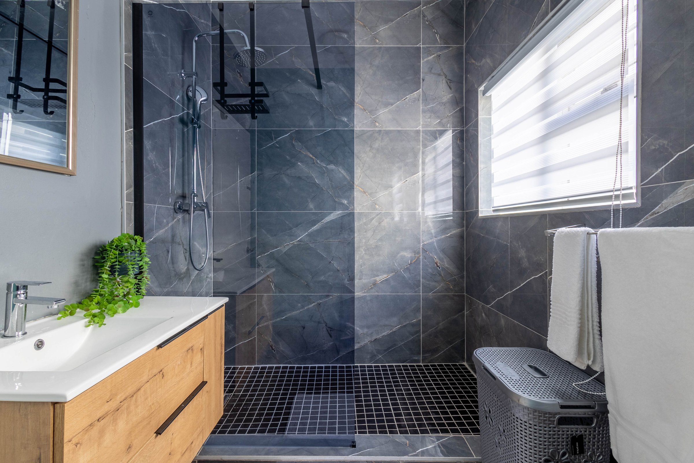 Modern bathroom with dark marble tiles, glass shower, wooden vanity, and a basket with towels.