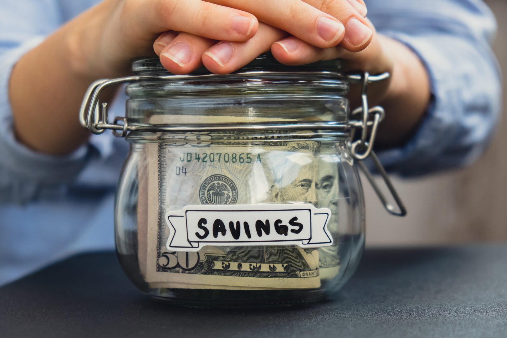 Unrecognizable woman holding Saving Money In Glass Jar filled with Dollars banknotes. SAVINGS transcription in front of jar. Managing personal finances extra income for future insecurity background