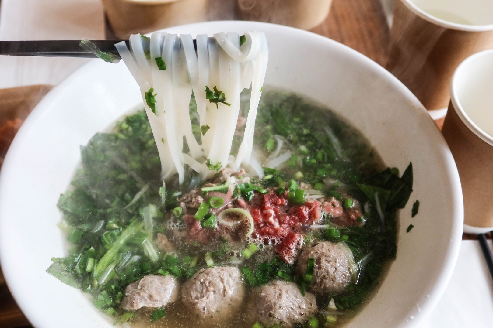 Vietnamese beef pho rice noodles with beef ball and thinly sliced raw beef added to a steaming bowl of savory broth. Chopsticks holding the rice noodles.
