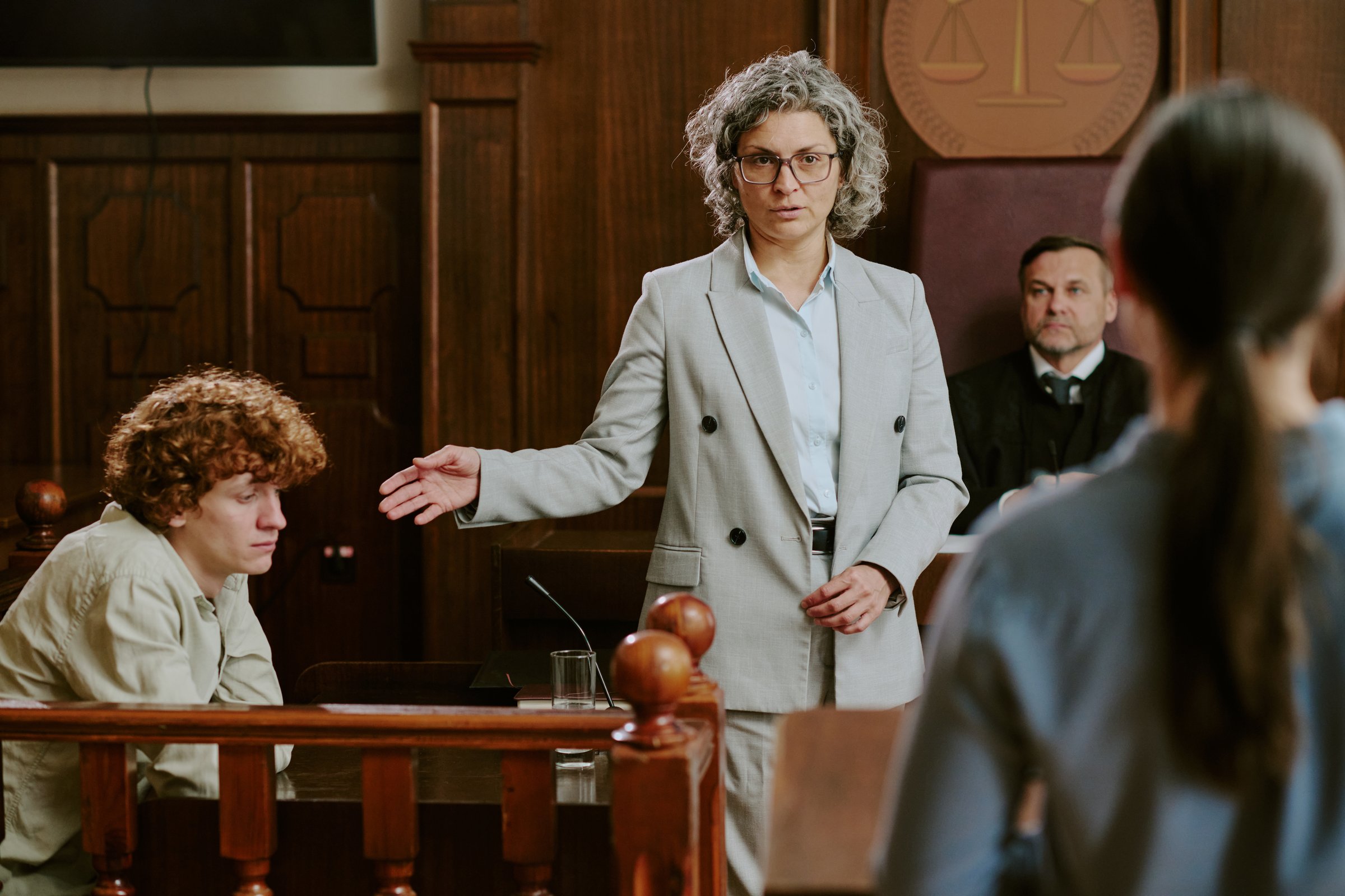 Middle aged Caucasian woman gesturing while standing in courtroom near seated teenage Caucasian boy, judge and another visible in background during legal proceeding
