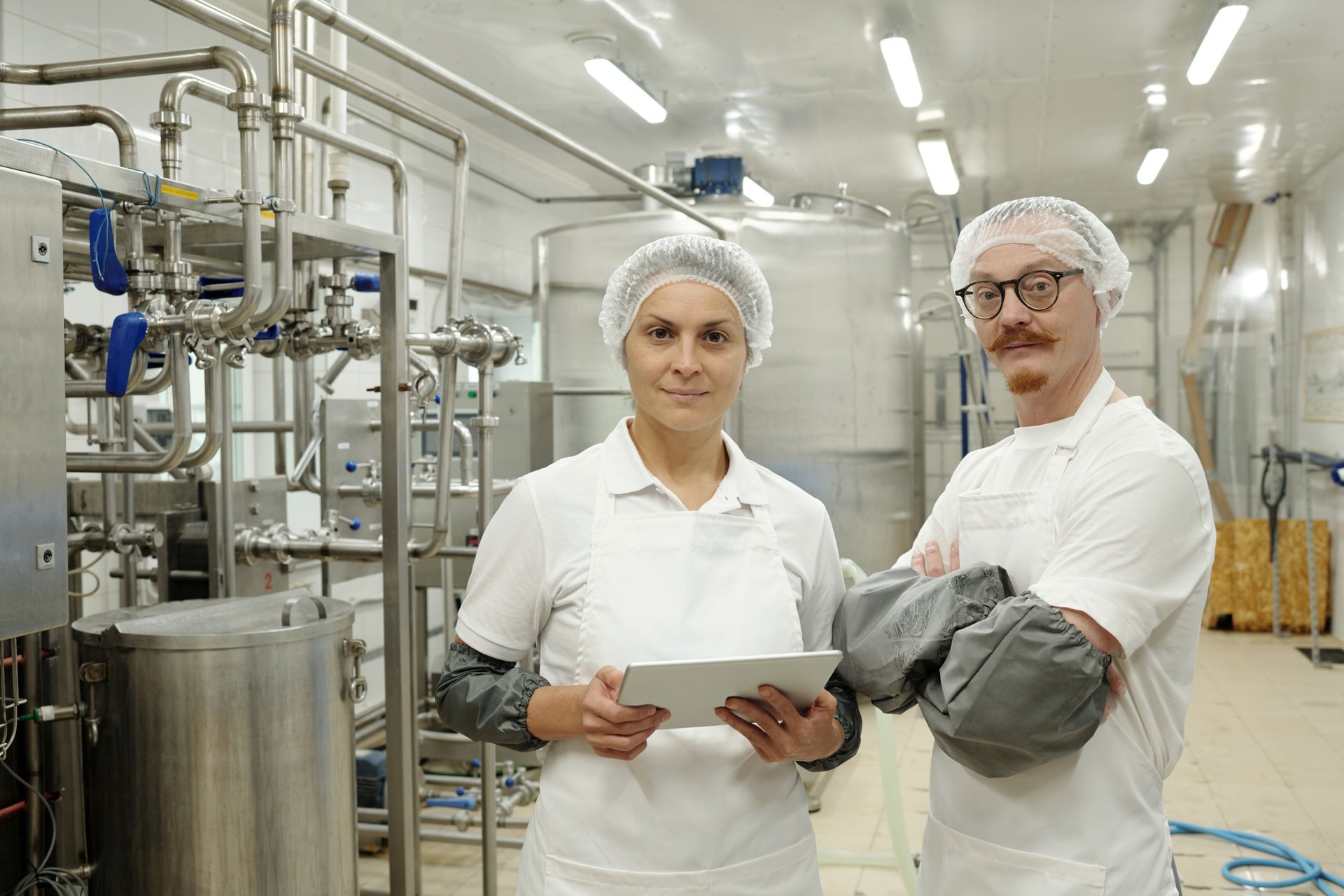 Inspecting equipment in food processing facility while wearing uniforms and hairnets. Worker holding tablet and technician looking at camera, ensuring cleanliness and safety standards