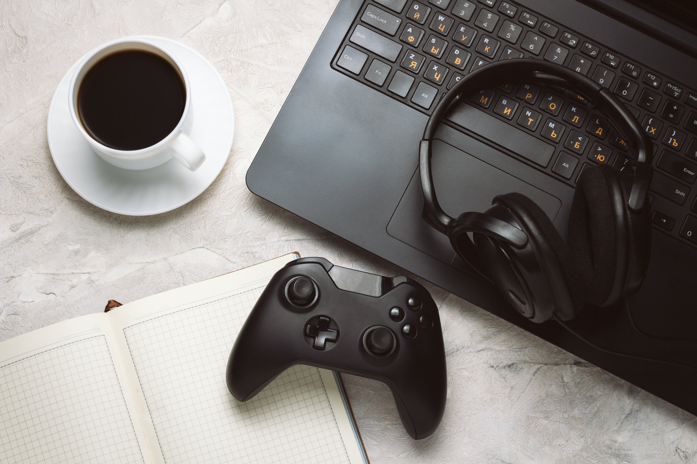 Top view of Gamepad, Laptop and Headphones, Cup with Coffee and Notebook on a White table with Сopy space. Gamers background concept.