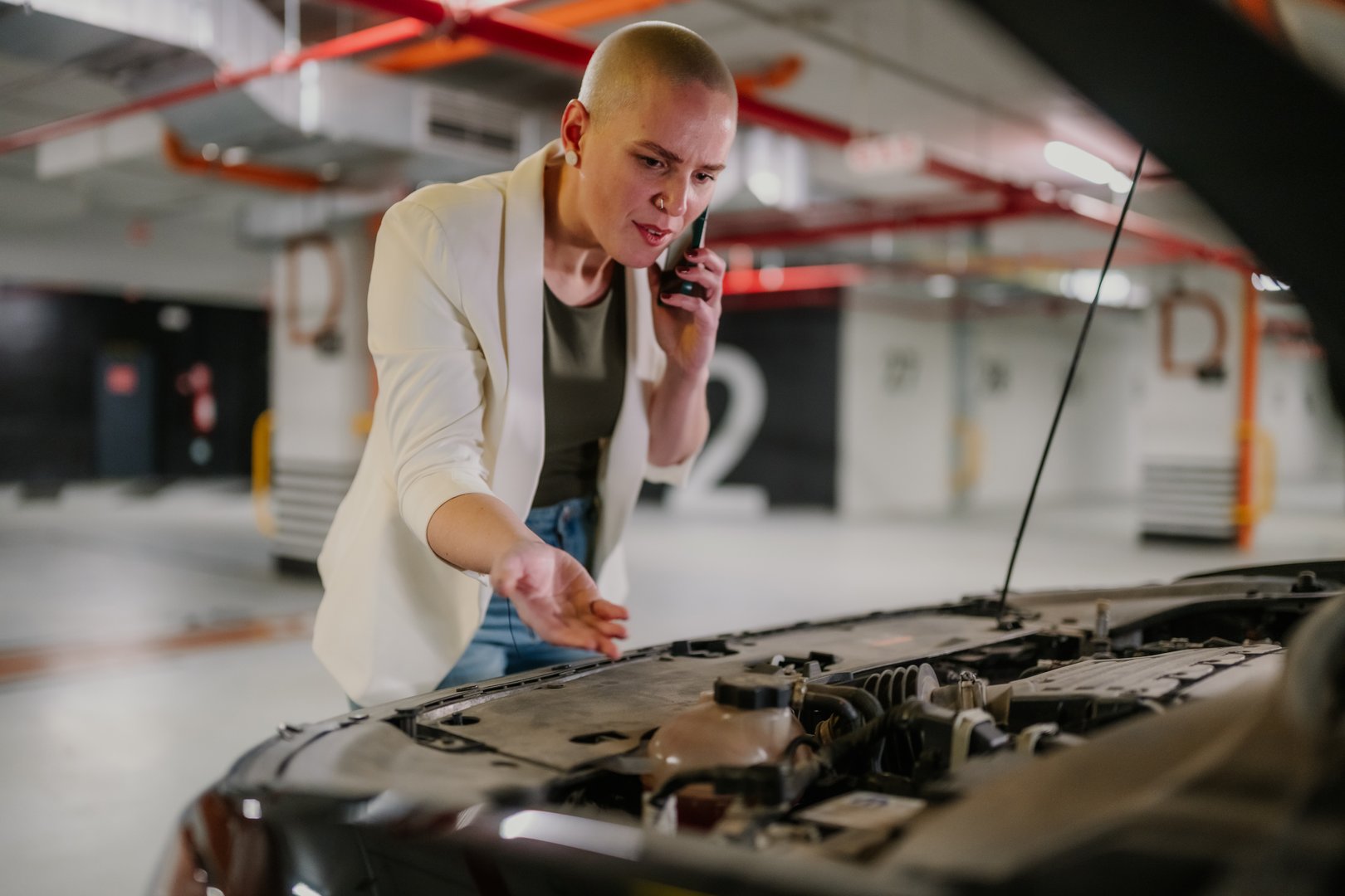 Young adult woman talking on smartphone while looking at open car hood, experiencing trouble with vehicle engine in an underground parking garage