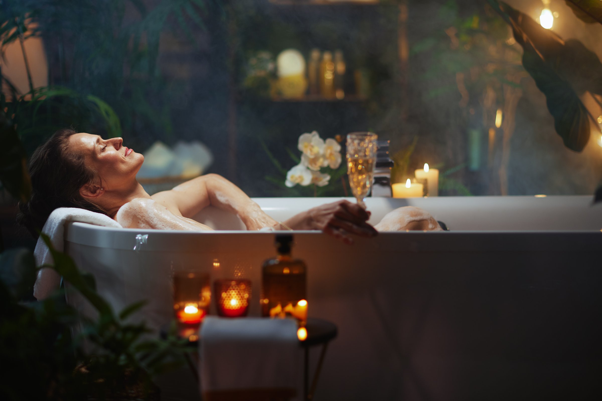 A middle-aged woman with her hair pulled back relaxes in a bubbly bathtub, holding a champagne glass. Lit candles, lush greenery, and soft, warm lighting create a luxurious, spa-like atmosphere for self-care and indulgence.