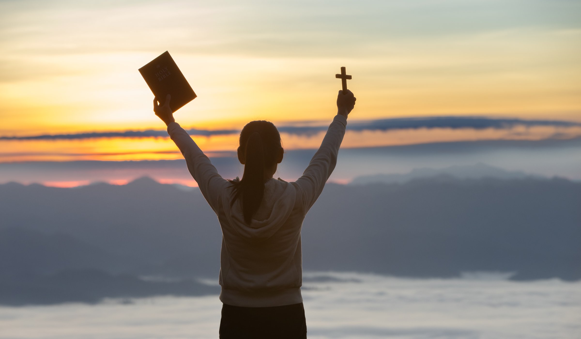 Silhouette of woman praying while holding Bible and cross, Pray in the Morning , Woman praying with hands together on the Sunrise background.