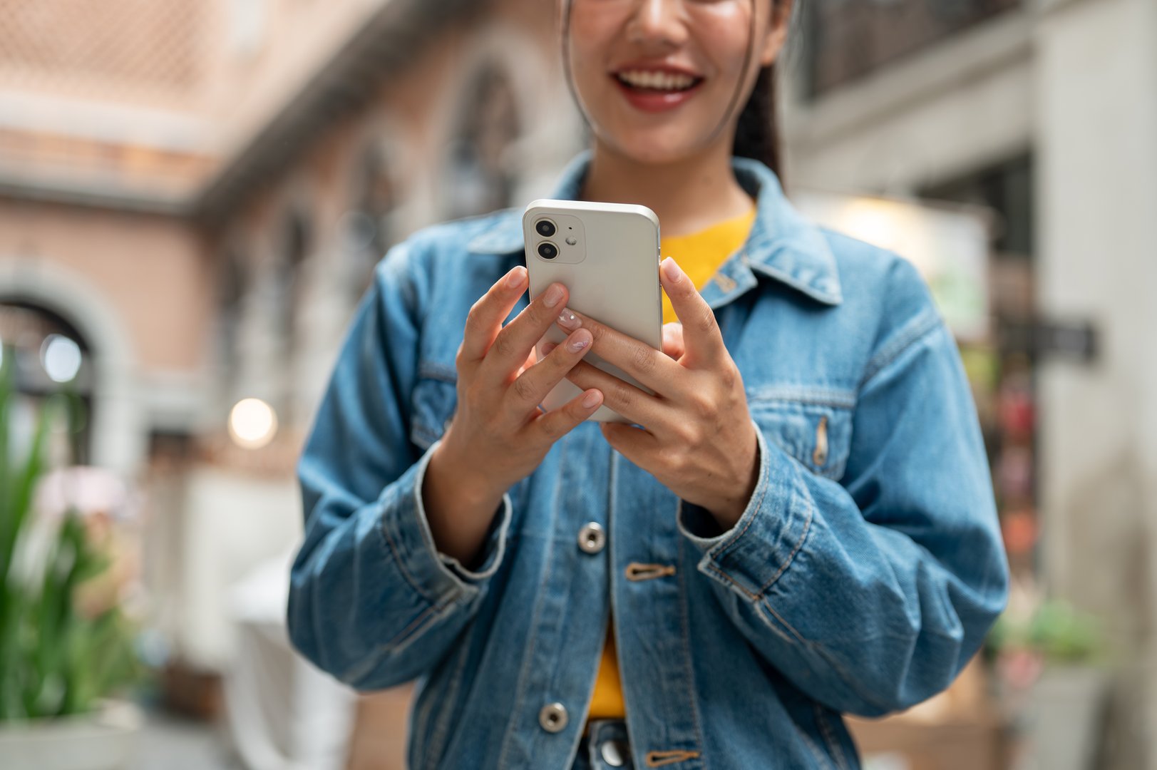 A cropped of a beautiful Asian woman in a denim jacket is standing outdoors with stores in the background, using her smartphone to read text or reply to a chat. people, lifestyles, wireless technology