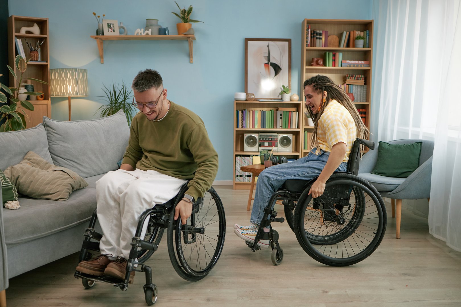 Young adult man and woman with disability sitting in wheelchairs laughing together in living room enjoying social interaction
