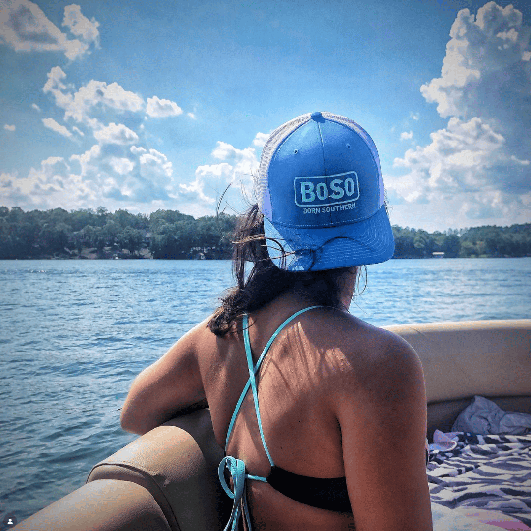 Woman in a bikini and blue cap with BOSO logo, seated on a boat, looks out at a lake under a partly cloudy sky.