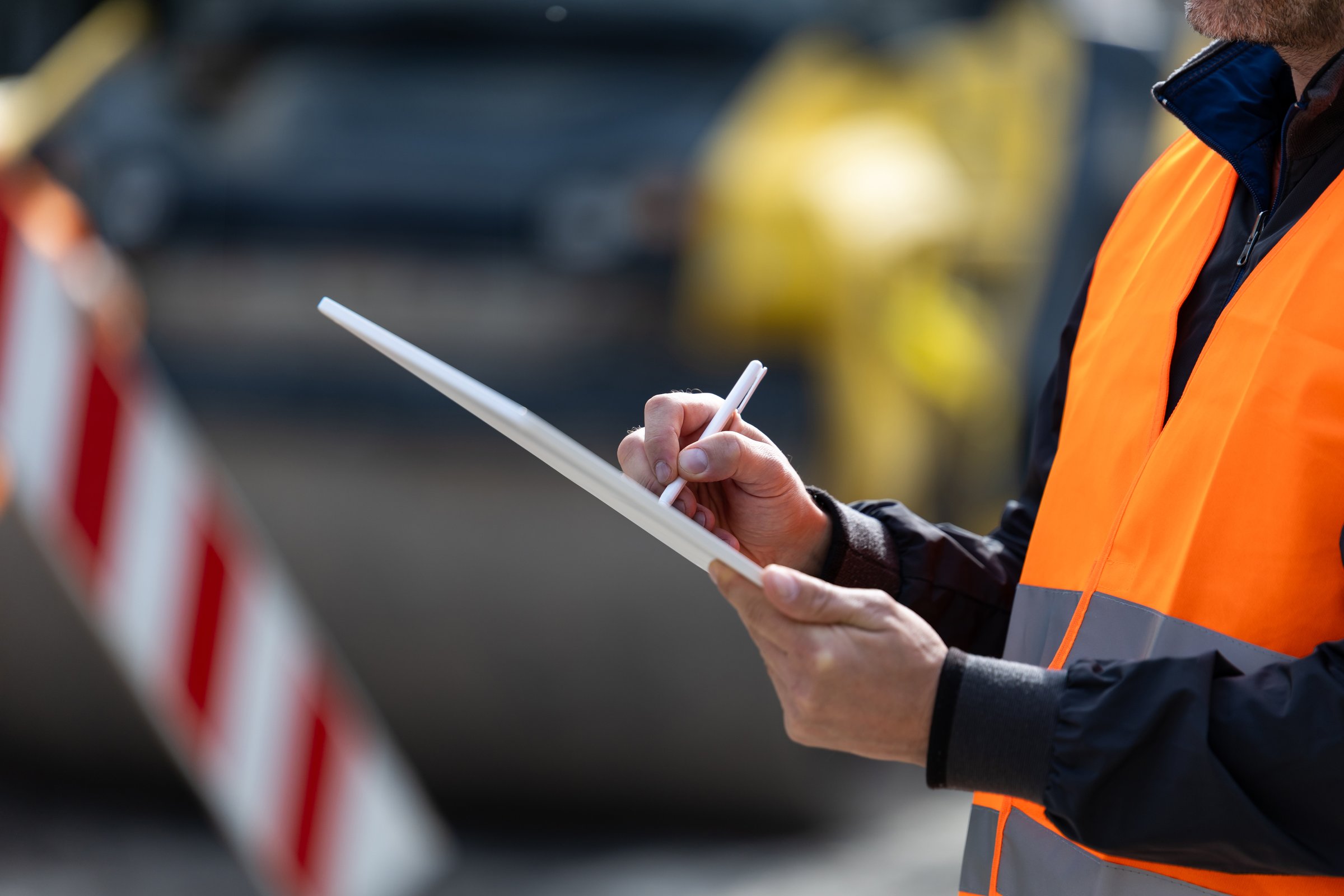 A construction worker, dressed in a safety vest, is focused on taking notes while observing ongoing activities at a road construction site under clear skies.