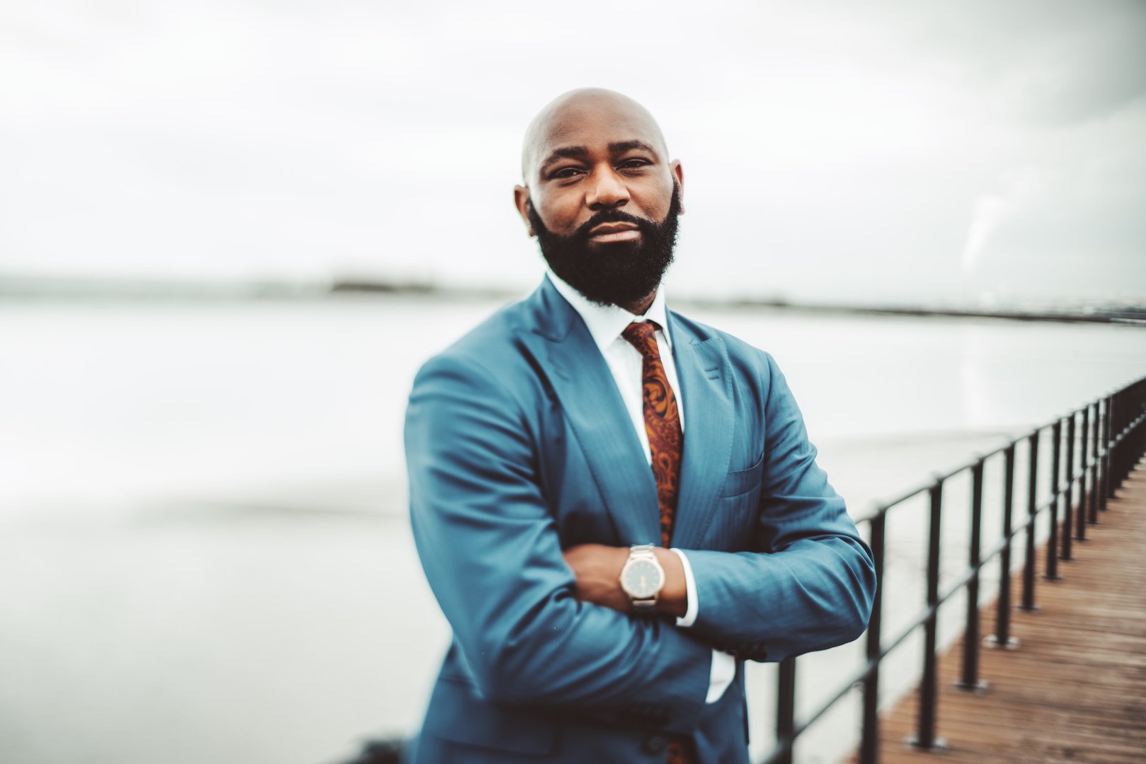 A true tilt-shift portrait of a successful adult African bald bearded man in a blue suit with tie and watch standing with hands crossed on an embankment near facing on an overcast day, selective focus
