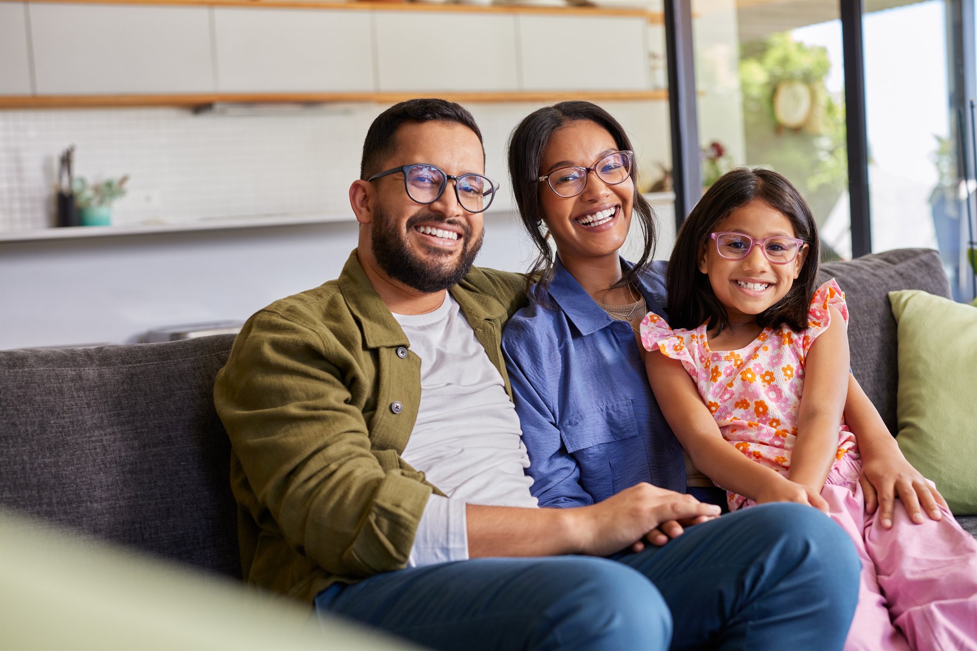 Portrait of happy indian family wearing spectacles at home while relaxing on sofa. Handsome father and beautiful mother sitting with daughter on couch wearing eyeglasses with blue block lens. Happy middle eastern family smiling and embracing while bonding together at home sitting on sofa.