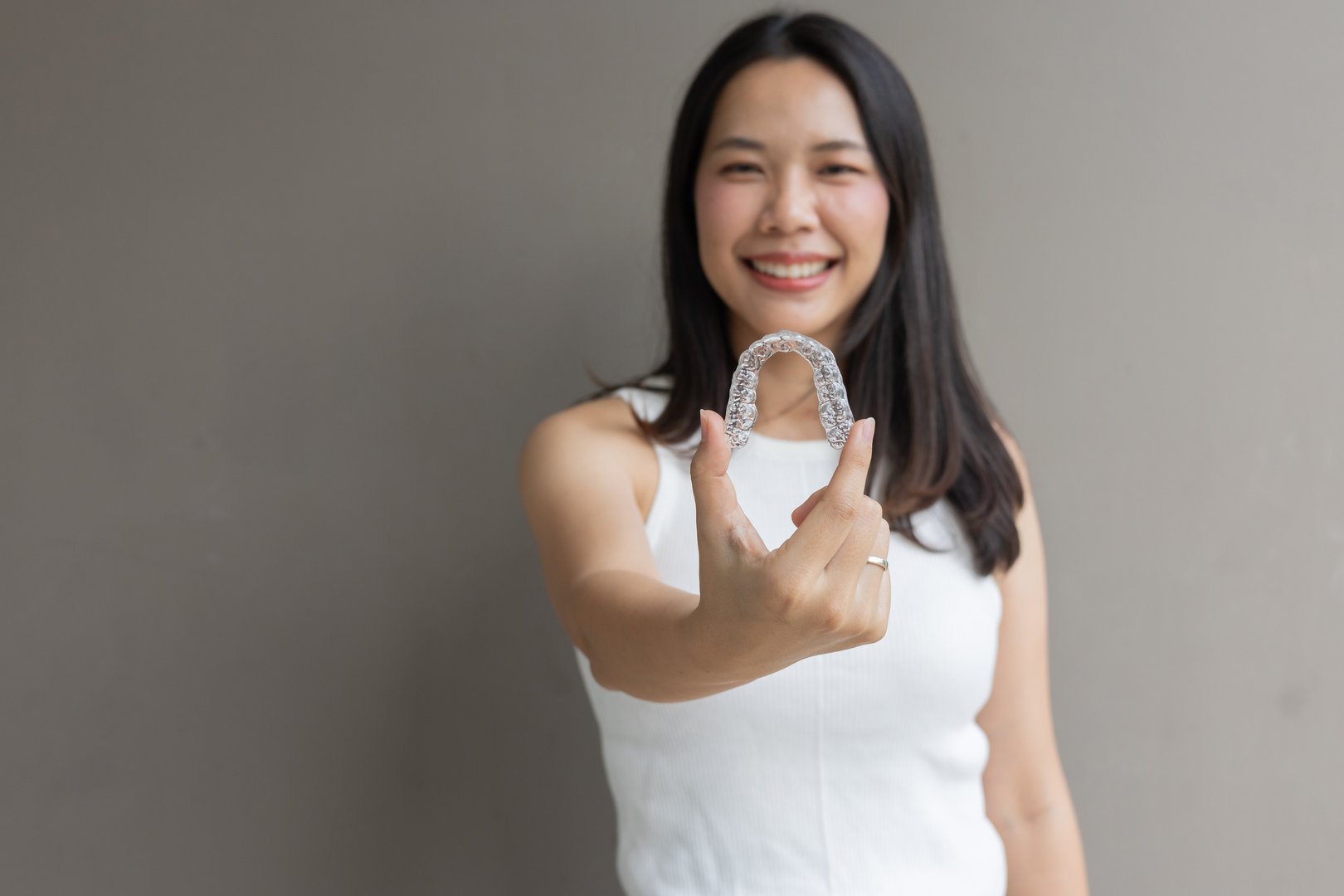 close up young asian beautiful woman smiling with hand holding dental aligner retainer (invisible) on grey background at dental clinic for beautiful teeth treatment course concept