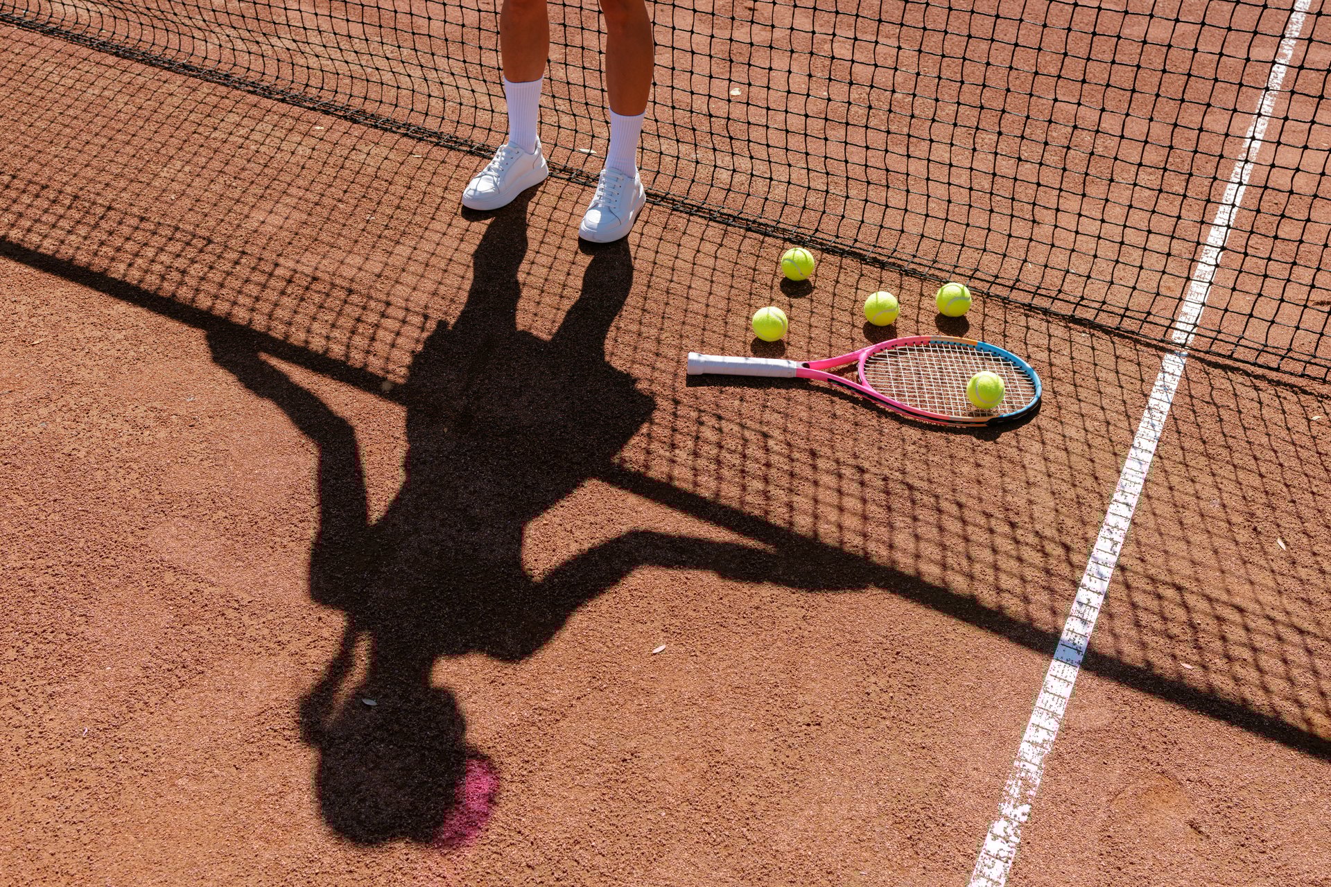 Athlete shadow on clay tennis court with racket and balls near net in bright sunlight, minimal sports background with copy space