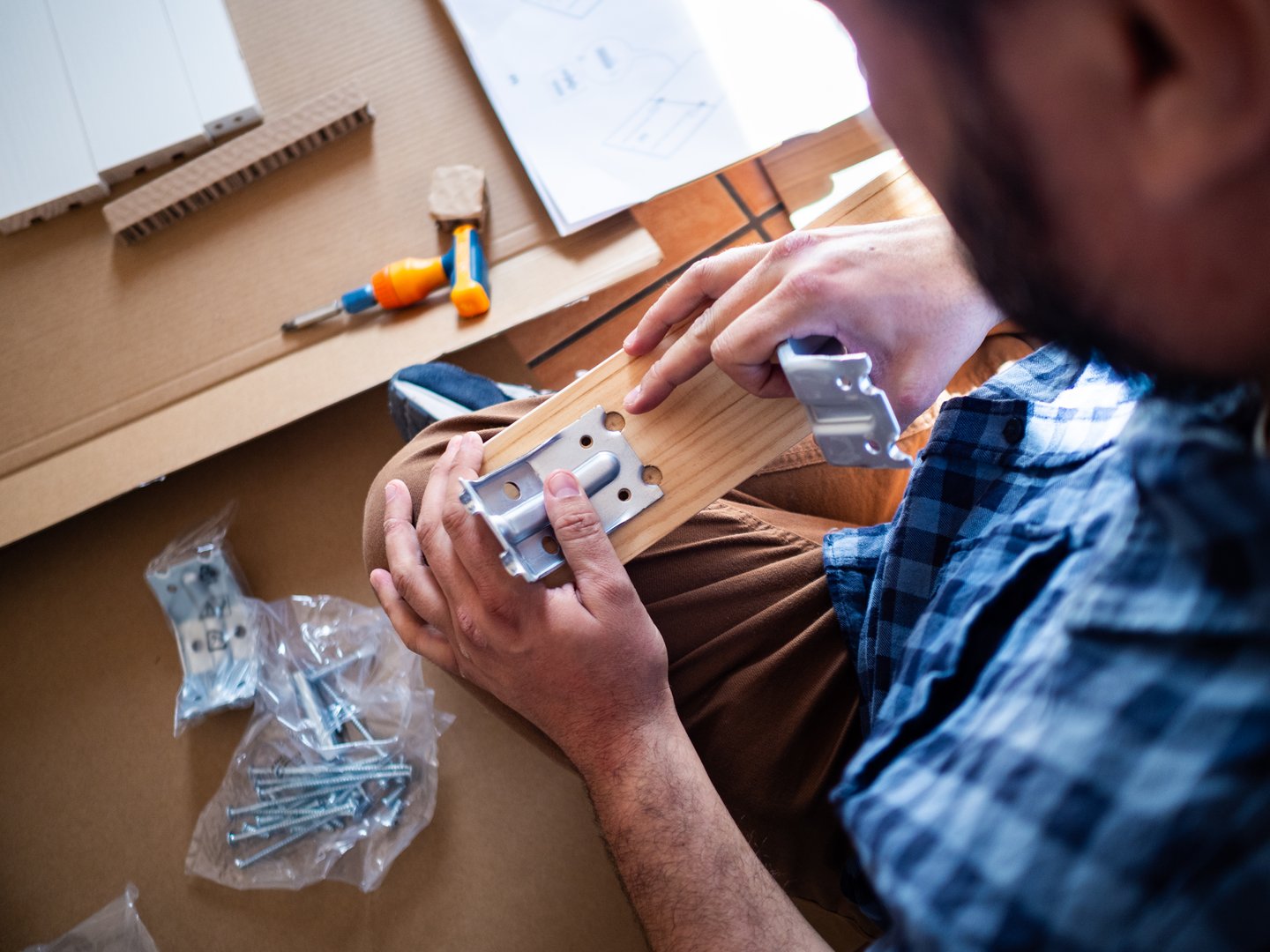 A person is assembling furniture, holding a wooden panel and aligning metal brackets. Various tools and screws are scattered around on the floor, creating a focused atmosphere of home improvement.