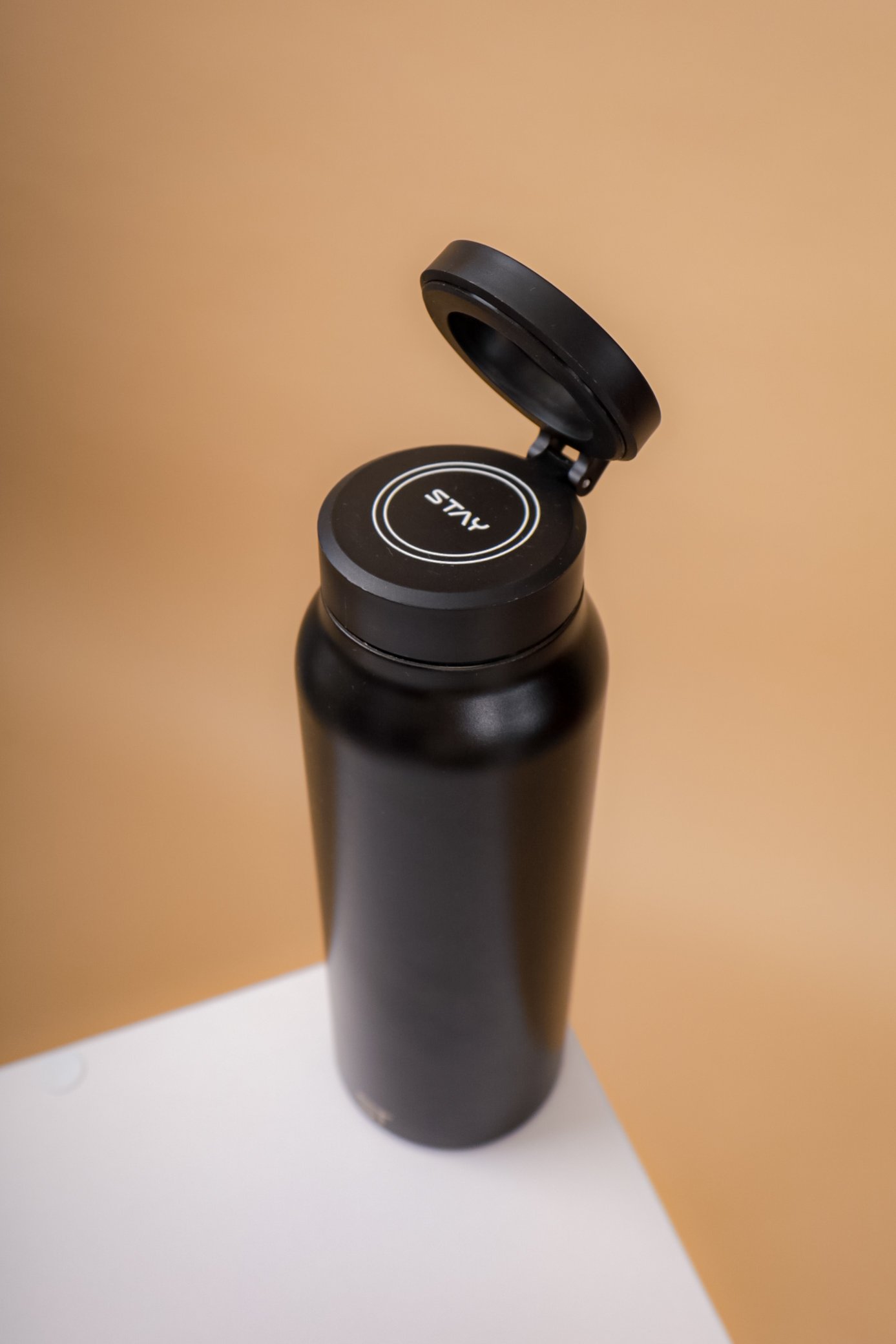 Black water bottle with a flip-open lid on a white surface against a beige background.