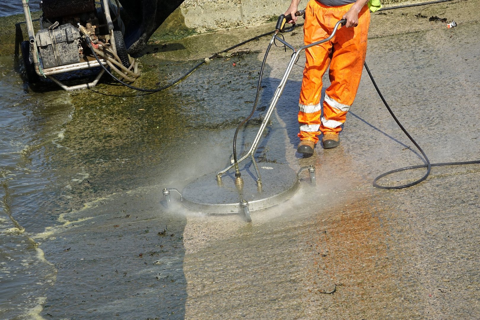 Plymouth England. The Cattewater. A man dressed in orange overalls cleaning seaweed off a concrete boat slipway using a water jet from a power washer drawing water from the sea. Cleaned area clearly defined. River Plym and dicks in background.