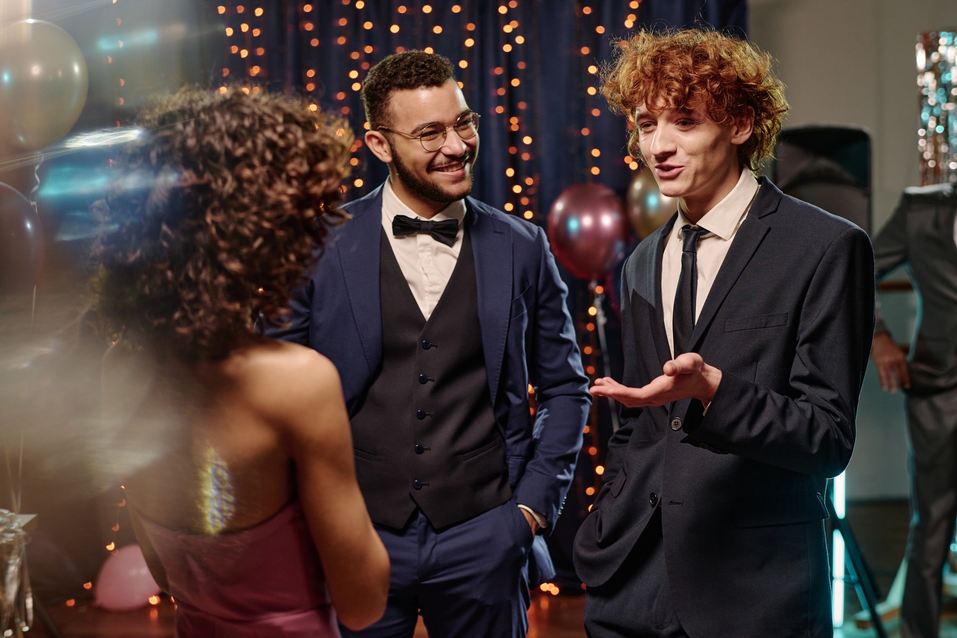 Teen boys wearing formal suits standing together, smiling and conversing with young woman at festive event with balloons and string lights in background