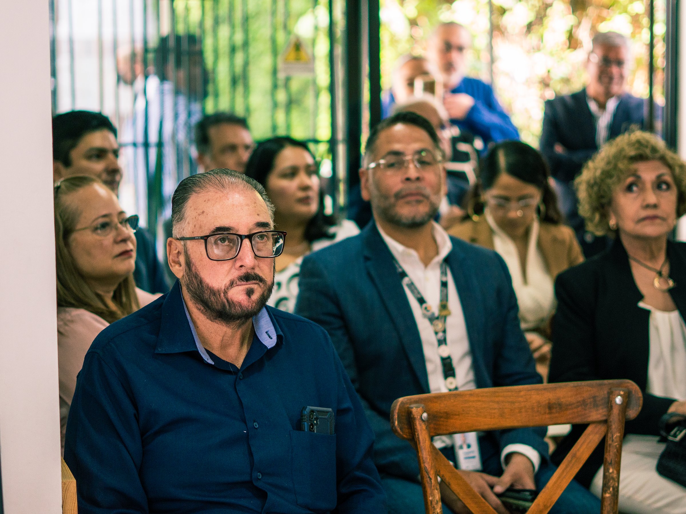 A group of people sitting attentively in a room, focusing on a presentation or discussion, with a man in glasses in the foreground.