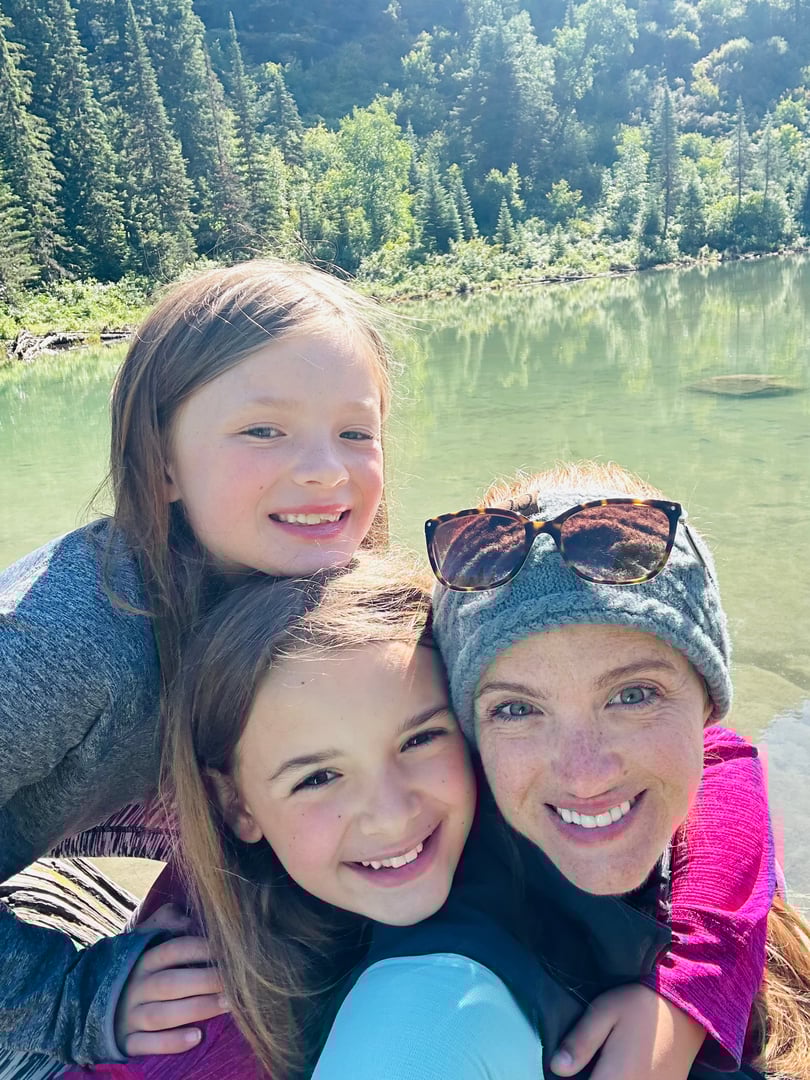 A woman with two children smiling by a serene lake and forest background, all wearing casual outdoor clothing.