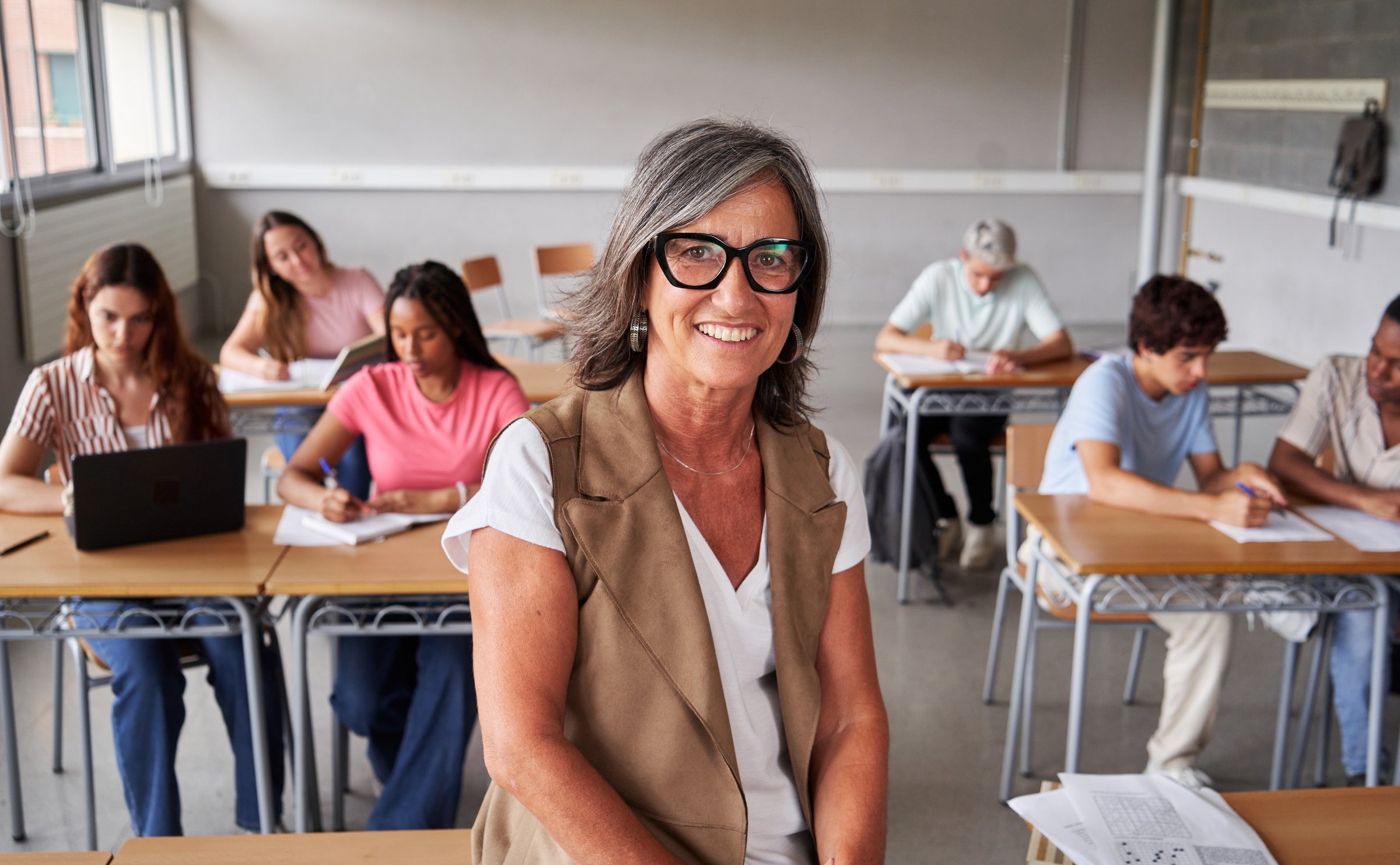 Portrait of a cheerful mature female teacher at classroom working, looking at camera smiling and confident while teenager students study in the background. Concept of professional occupation.
