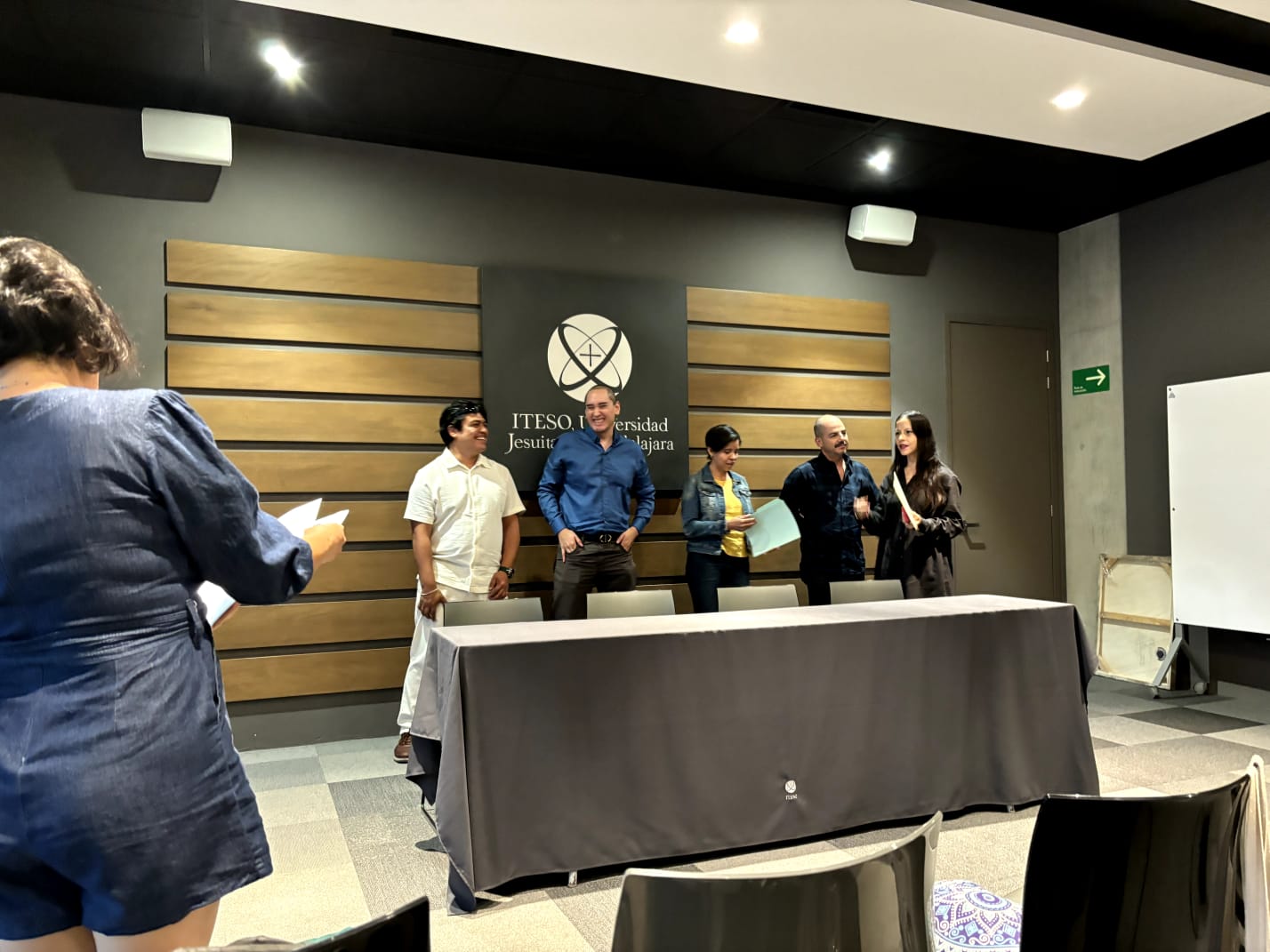 People standing and chatting in a conference room at ITESO Universidad, with a table and chairs in the foreground.