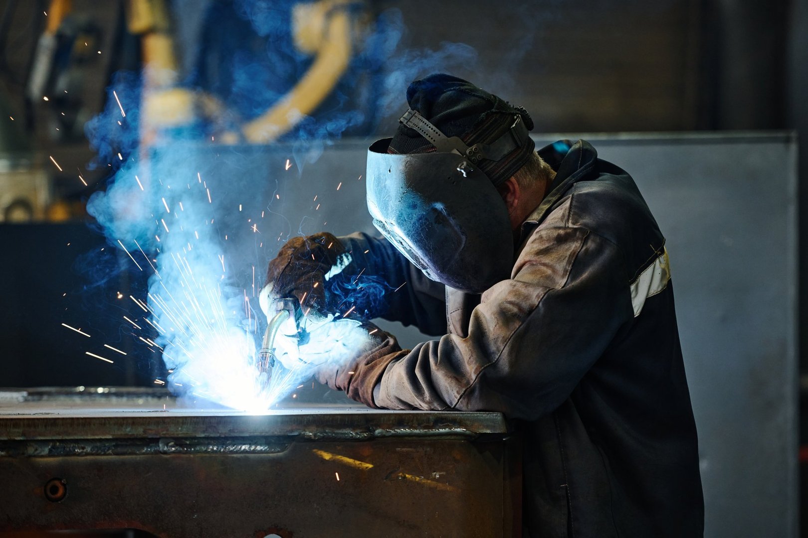 Skilled worker wearing protective gear and helmet welding metal piece, creating bright sparks in industrial setting. Precision and technique evident in process