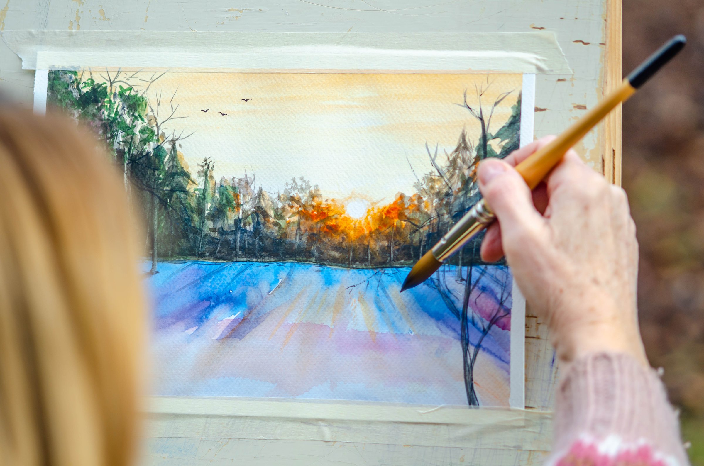 Senior woman painting a watercolor outdoors in winter, capturing the serene beauty of the winter snowy landscape. She is focused on her artwork, surrounded by a calm, cold environment with few snow-covered areas. The scene evokes a sense of peaceful creativity and winter solitude, showcasing the artist’s dedication to her craft in the crisp, frosty air. Ideal for themes of aging, winter, art, and outdoor creativity.