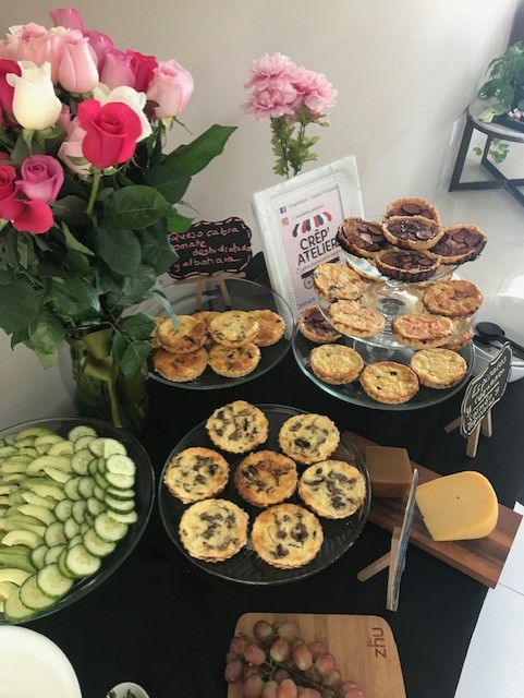 A buffet table with assorted quiches, cut cucumbers, roses, and a small cheese platter. Display includes a "Crep Atelier" sign.
