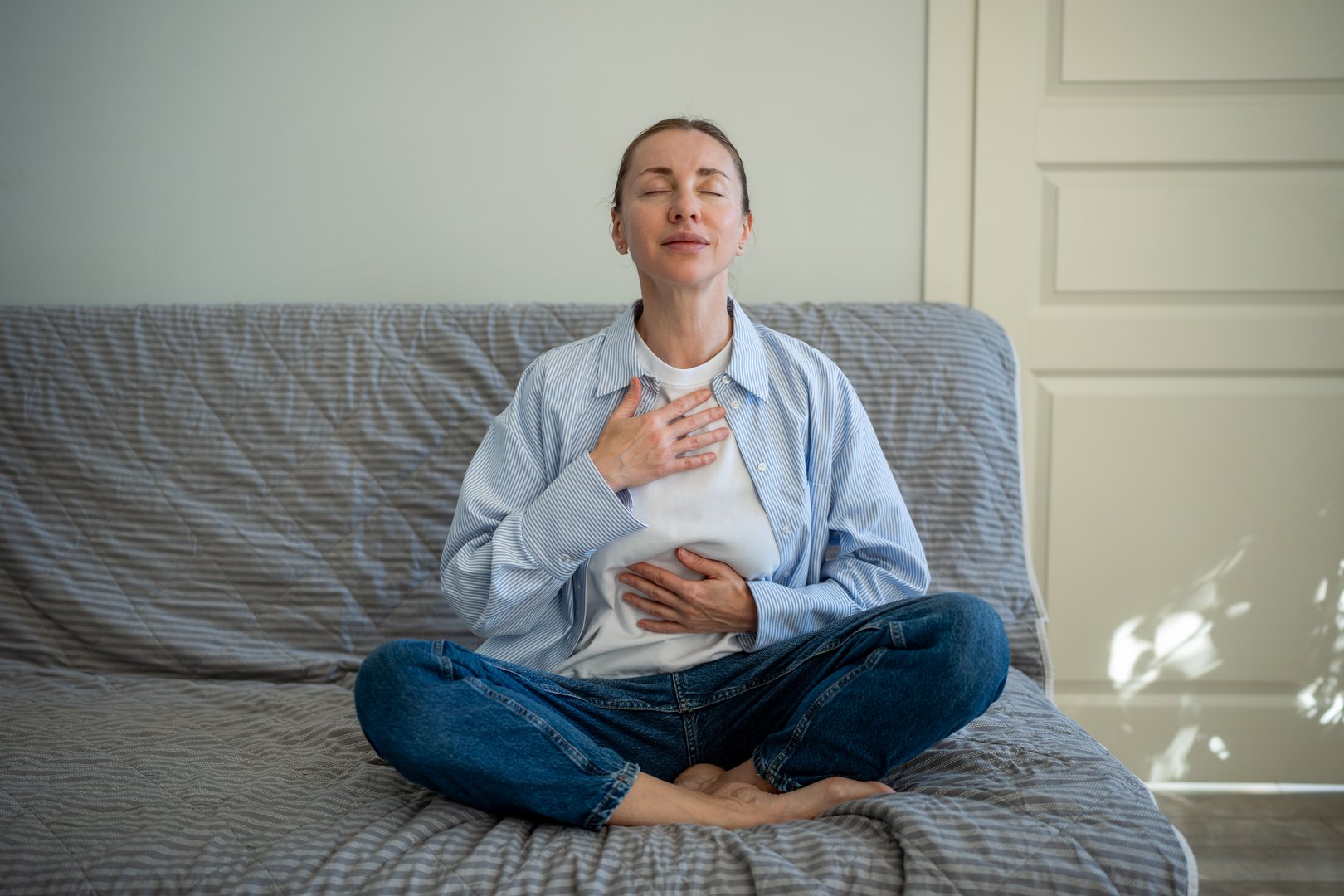 Calm woman sitting in lotus pose deep breathing during meditation at home. Self-help of middle aged female stress relief in daily routine reset mind with eyes closed, placing hands on chest, abdomen.