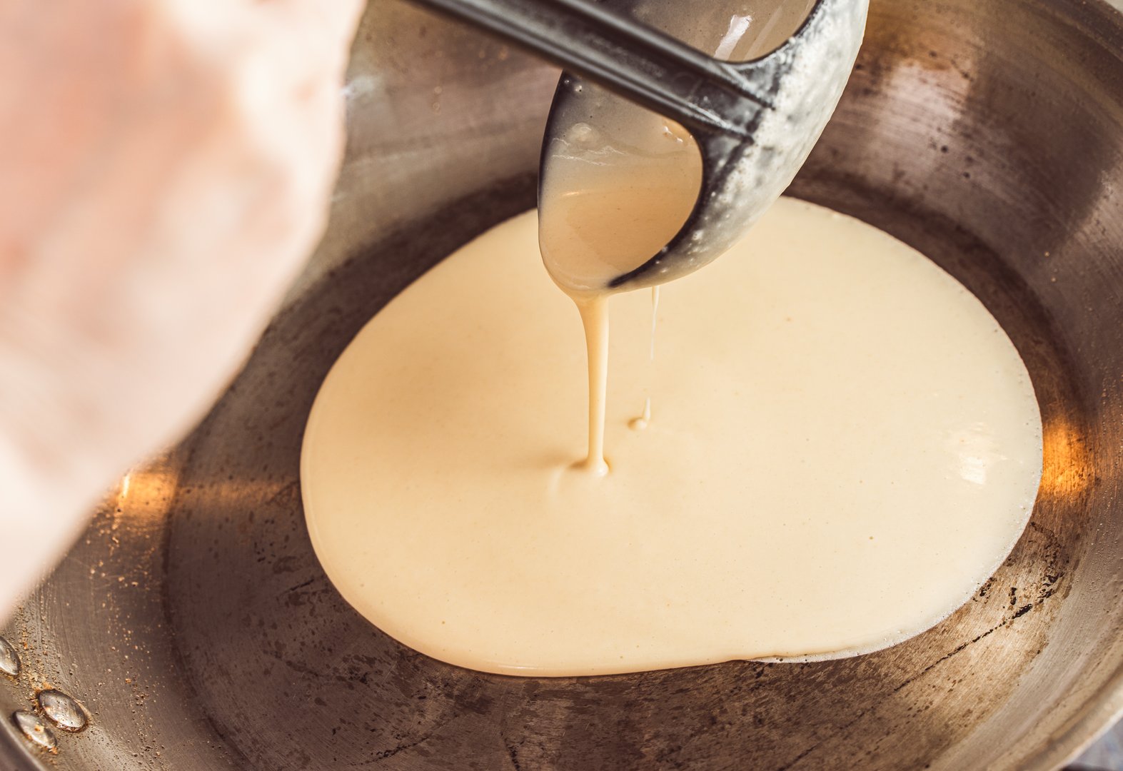 A woman's hand pours pancake batter into a hot pan. Recipe Crepe Suzette. Close-up, chosen focus. High quality photo