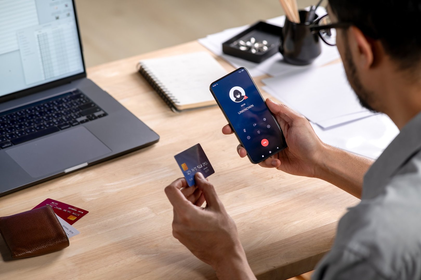A credit card user giving the credit card number to a scammer while holding a phone showing a scammer on the line