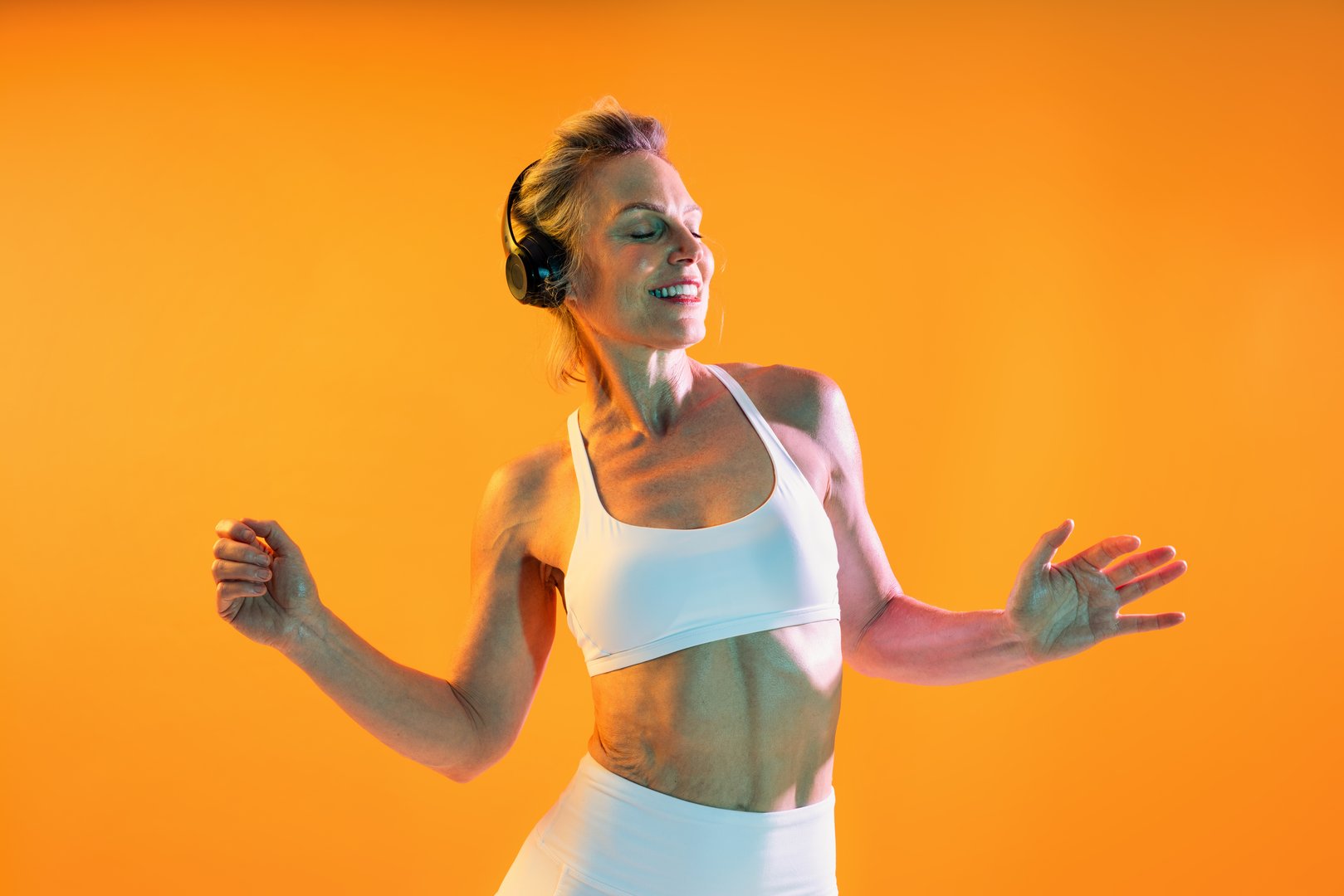 Beautiful mature sportive woman wearing sportswear doing workout - happy senior woman dancing and smiling with closed eyes in white sportswear on an orange background, enjoying music and fitness