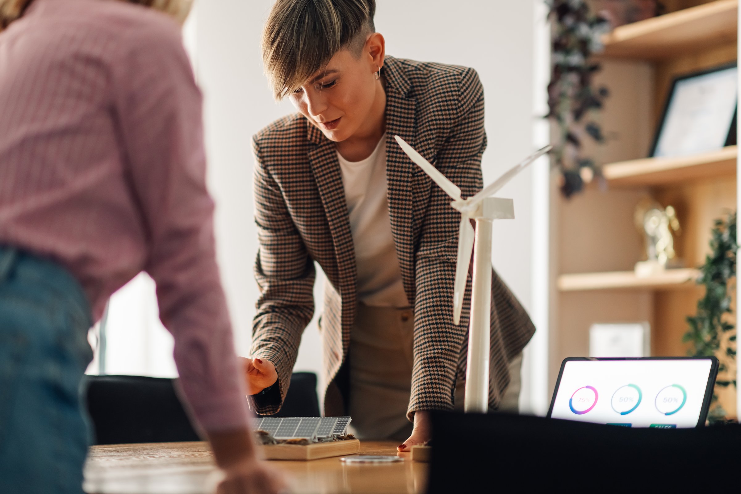 Two engineers are evaluating a project for renewable energy using miniature models of a wind turbine and a solar panel, discussing potential solutions for a sustainable future