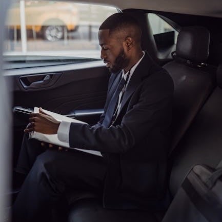 Man in a suit reviewing documents inside a car, seated with a focused expression, with a blurred cityscape outside the window.