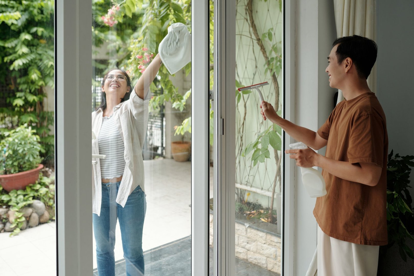 Two individuals cleaning windows together, one smiling and the other using a squeegee. Large glass doors reveal lush greenery outside, adding a serene and pleasant backdrop