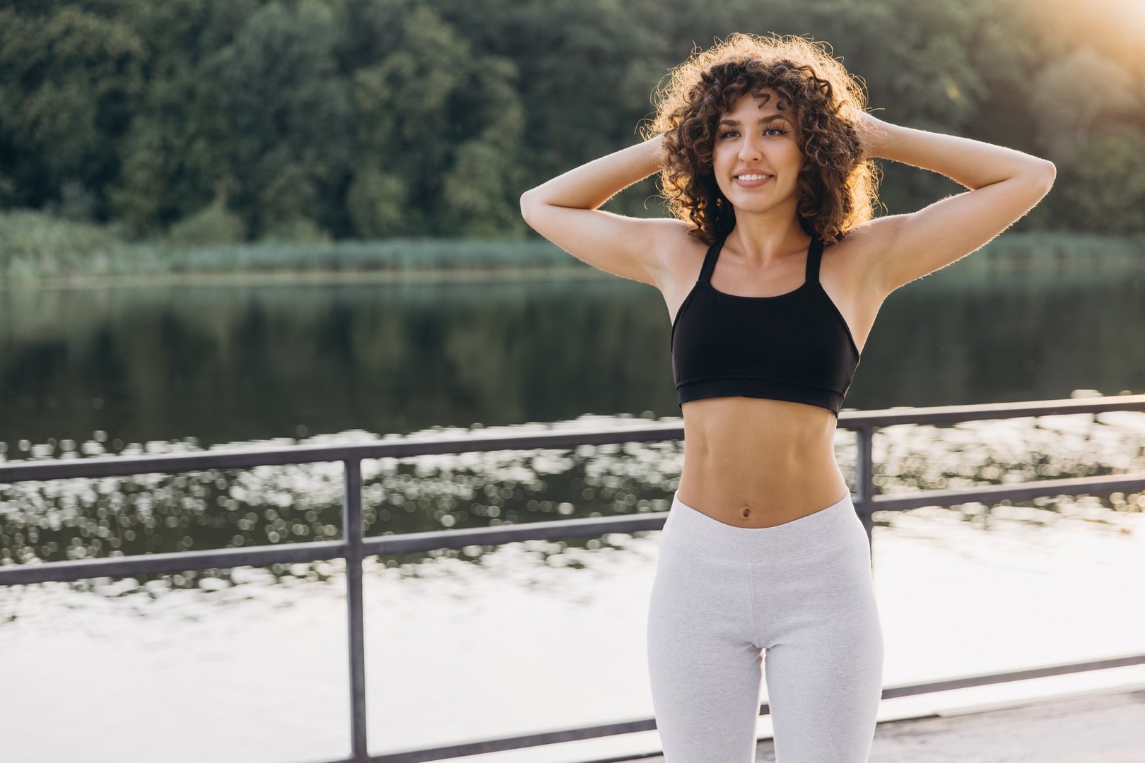 Curly haired sportswoman wearing black top and gray leggings stretching arms near lake during summer workout
