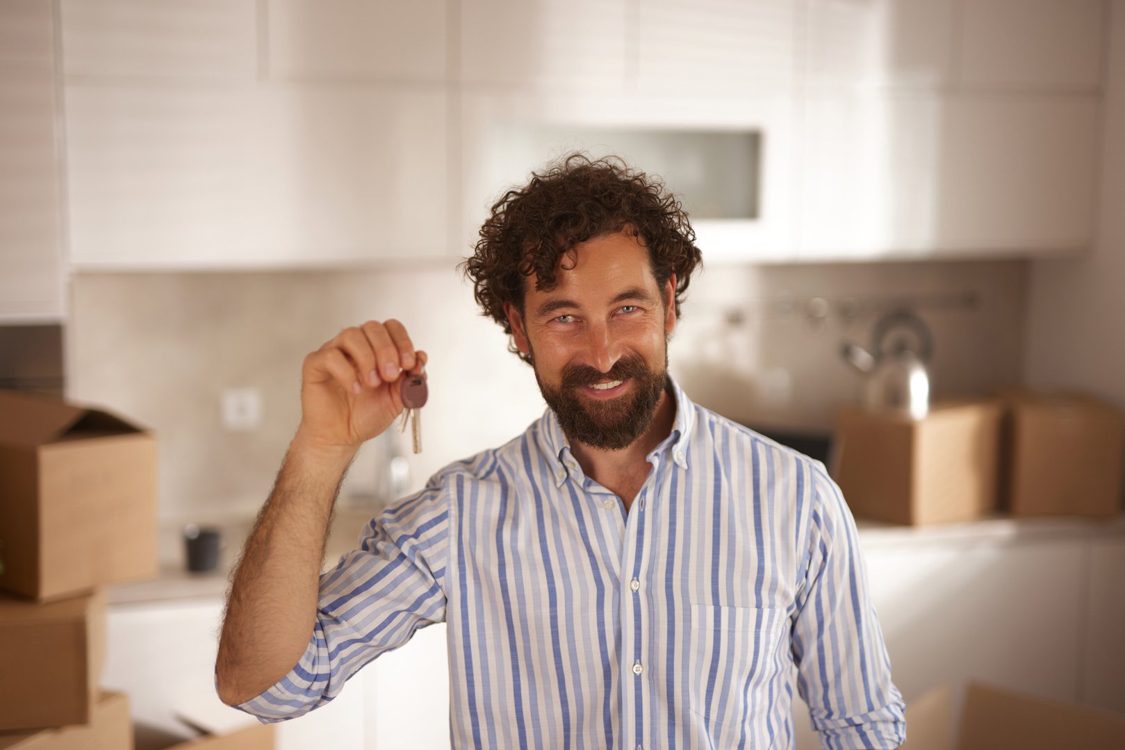 Mid adult man smiling and showing keys of his new house, with cardboard boxes in the background