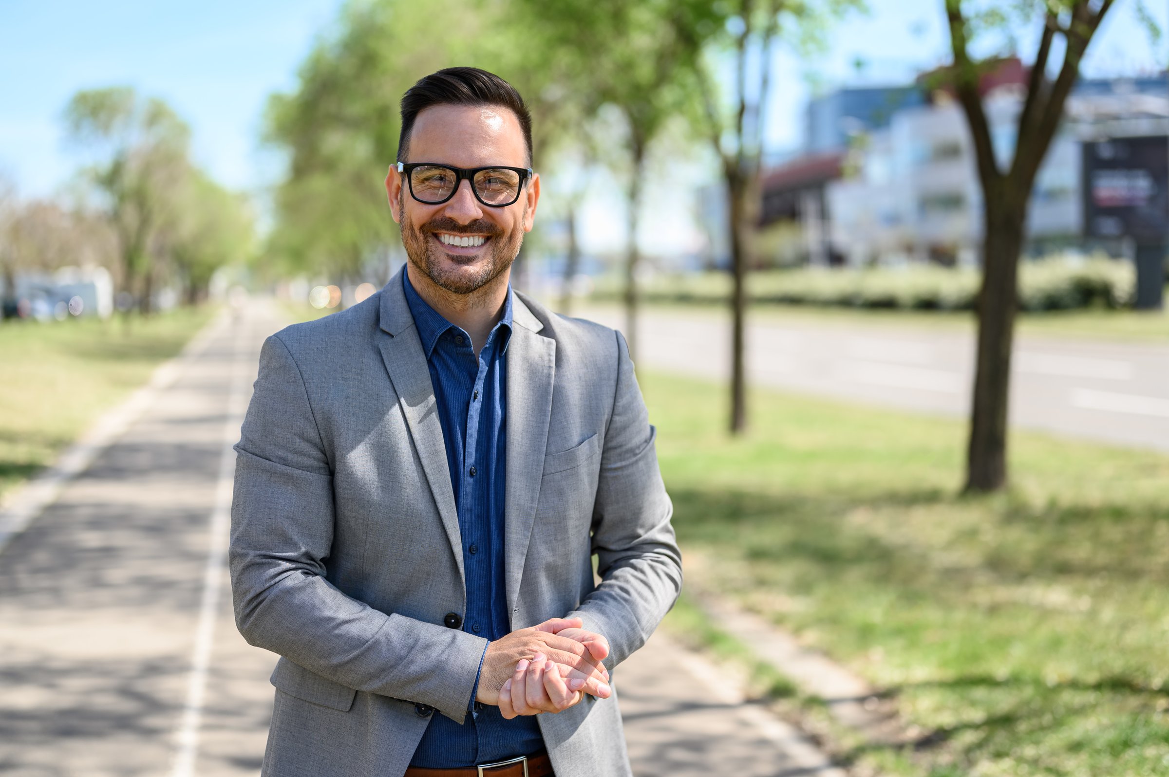 Portrait of contented young businessman with hands clasped smiling and standing on city street
