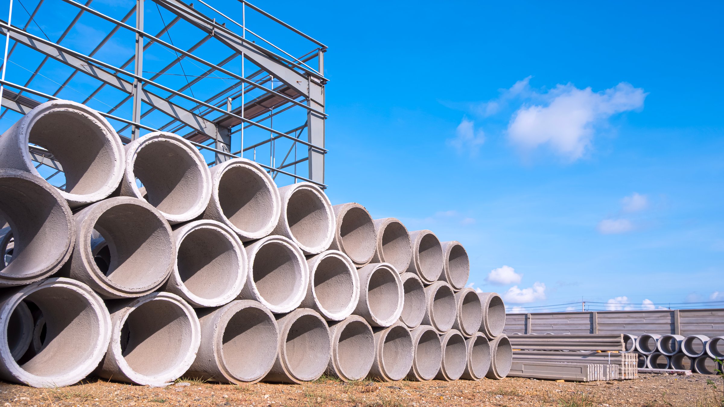 Stack of concrete drainage pipes on the ground in front of industrial building construction site against blue sky background, low angle and perspective side view