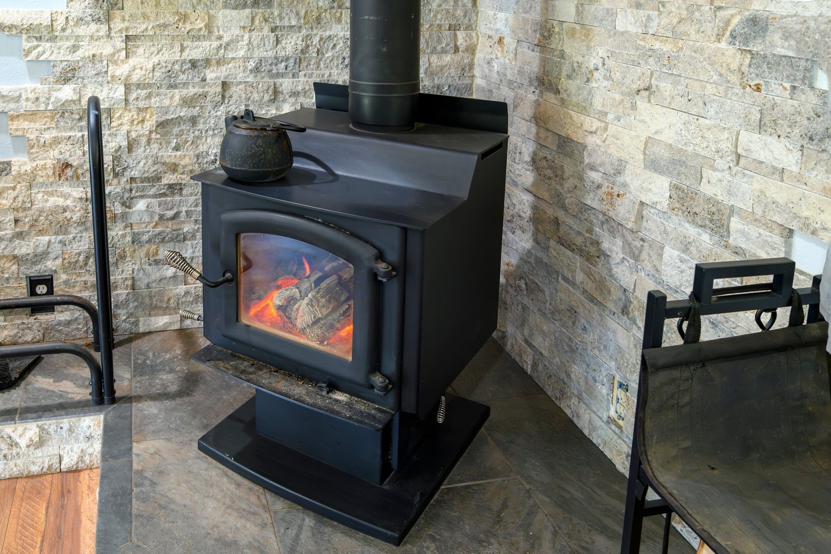 Burning logs seen through a window as a flaming wood stove gives off heat from a fire, with a stone rock fireplace surround behind it.