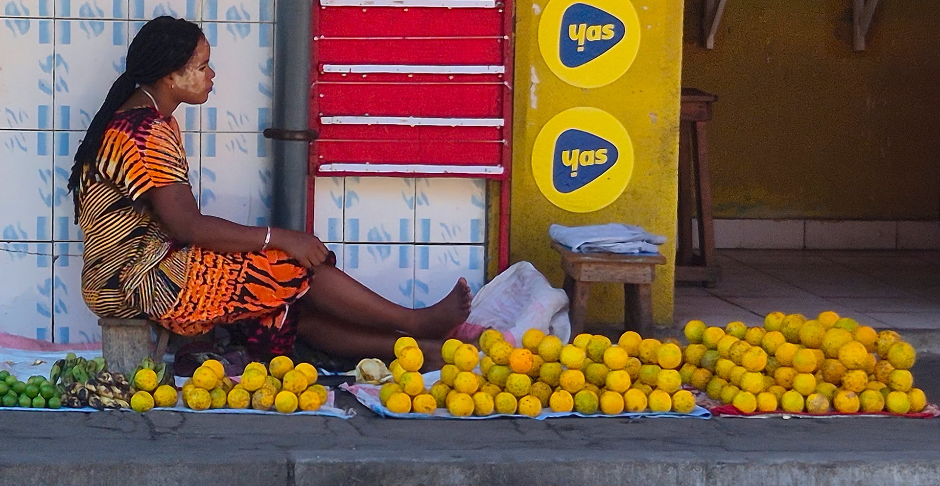 Morondava, Madagascar - August 14, 2025 : Local woman, sitting on the sidewalk, selling fruits. Her face is covered with a protective and decorative mask made of ground wood, called Masonjoany.
