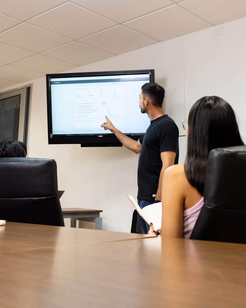 Man presenting data on a wall-mounted screen to seated colleagues in an office meeting.