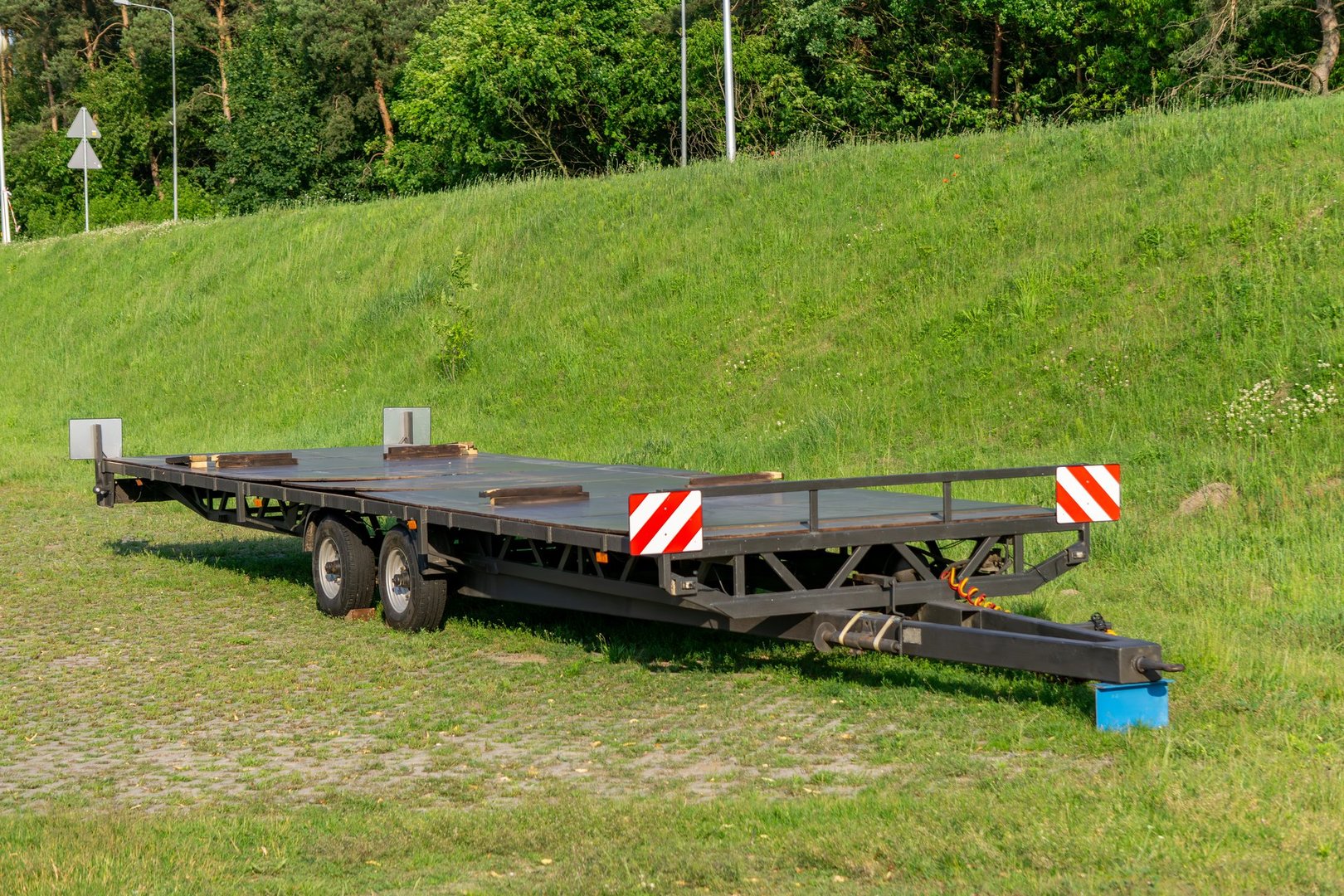 low-bed transport trailer with reflective markings and double axle, parked on grass near a road. Designed for carrying heavy equipment or machinery.