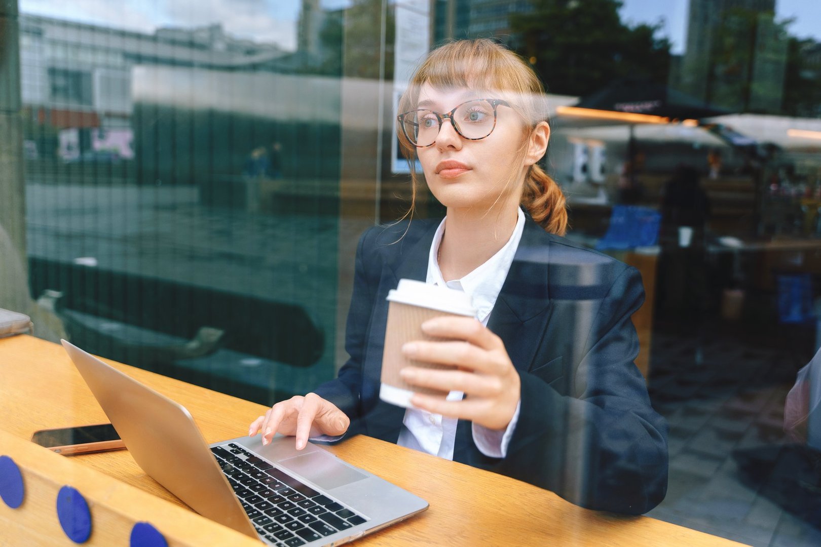 A focused young woman in a blazer types on her laptop while sipping coffee at a café, surrounded by a lively city backdrop on a busy morning.