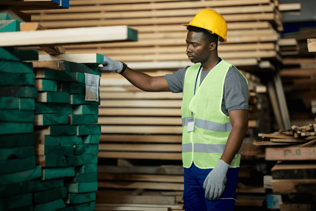 Worker in safety gear inspecting lumber stacks in a warehouse.