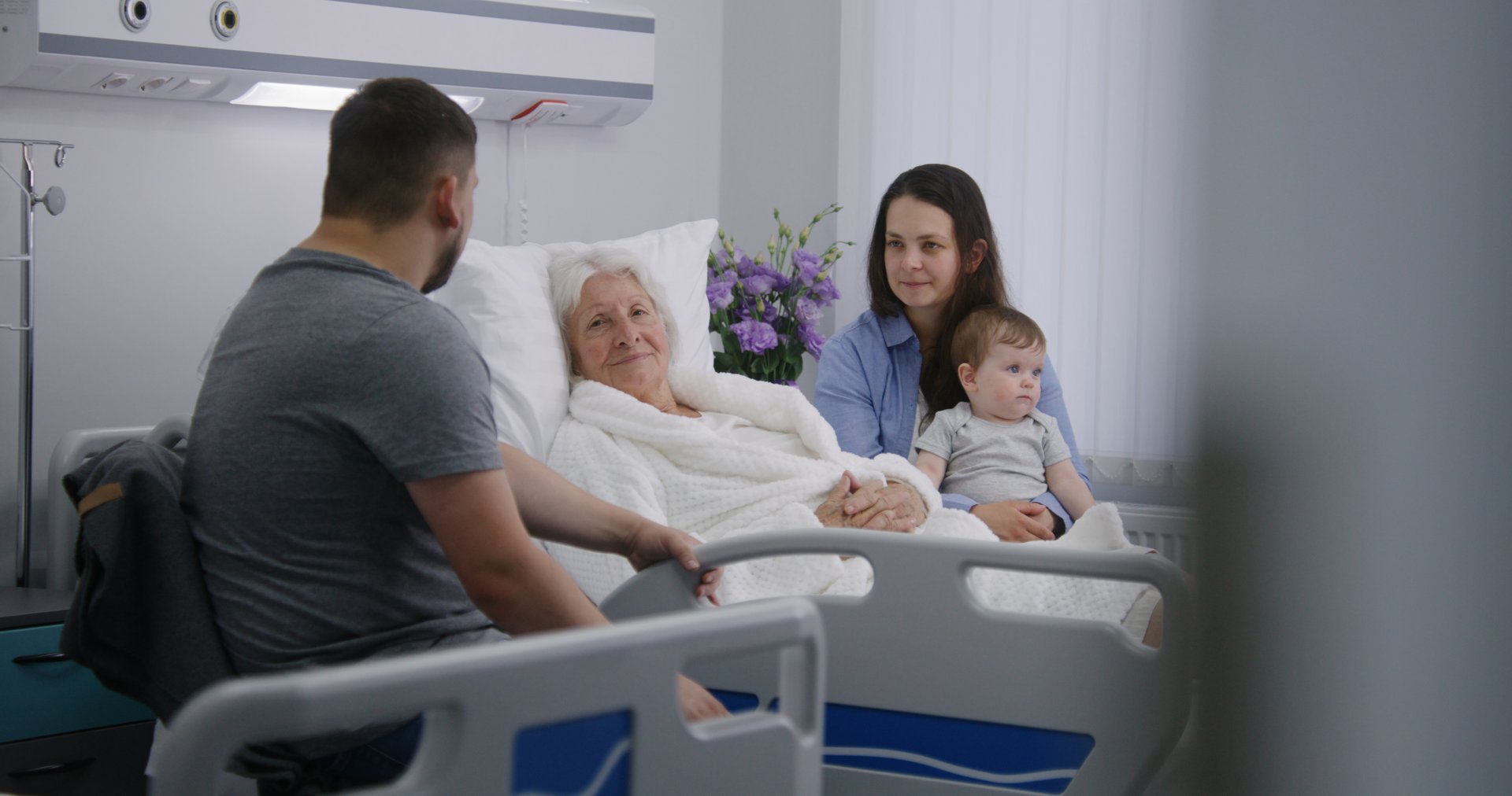 Elderly woman lying and resting in bed in bright hospital ward, plays with grandson. Loving family members support grandmother recovering after successful surgery. Modern medical facility or clinic.