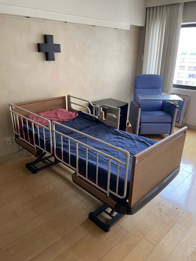 Hospital room with an adjustable bed, side table, armchair, and cross on the wall, bathed in natural light.
