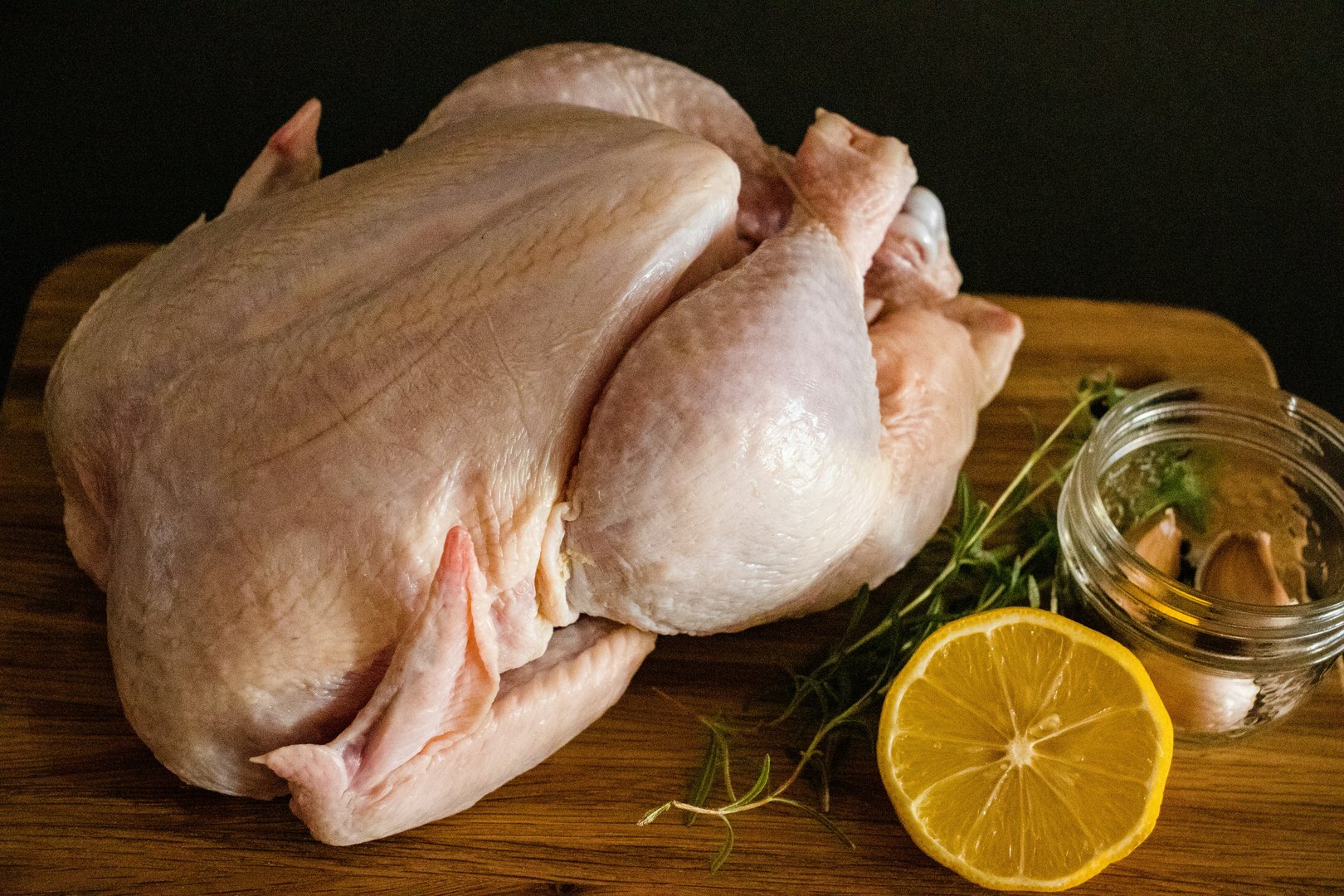 Whole raw chicken on a wooden board with a lemon slice, rosemary, and garlic cloves beside it.