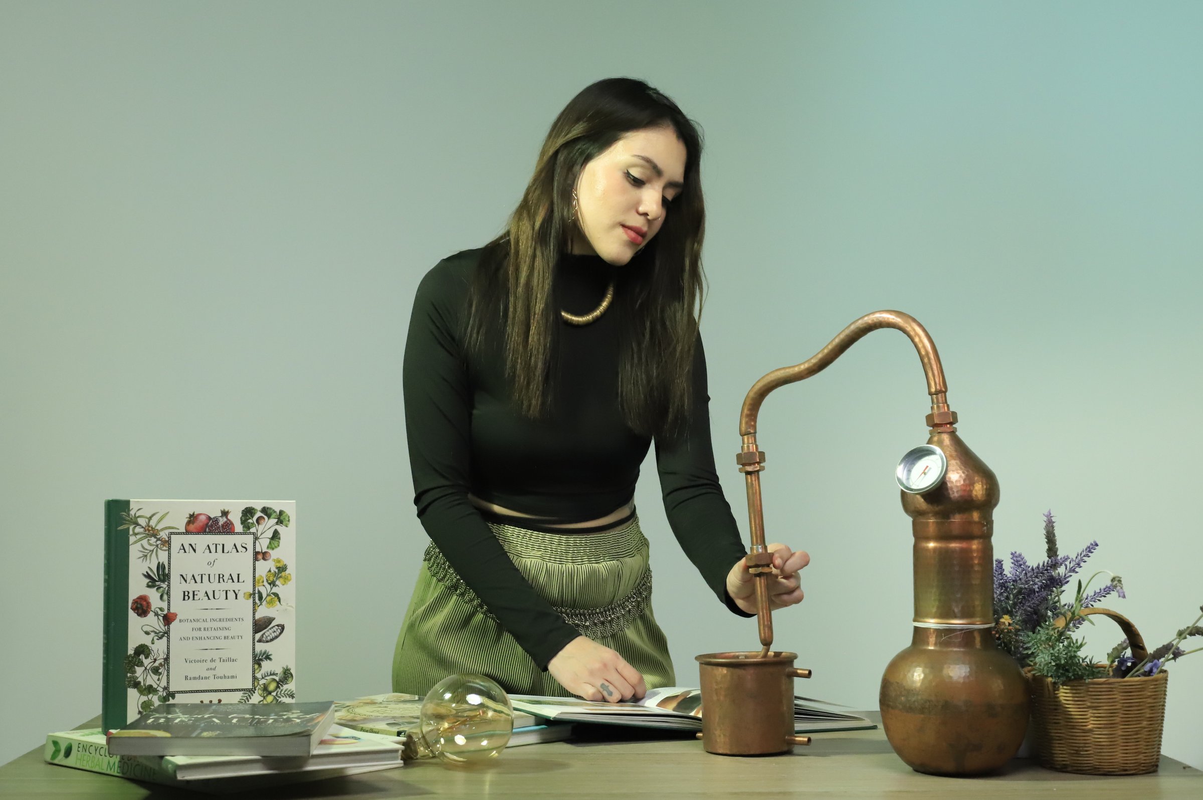 Woman in a black top using a distillation apparatus, with a book titled An Atlas of Natural Beauty and flowers on the table.