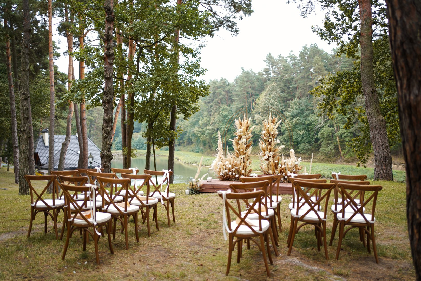 Rows of wooden chairs facing wedding arch with flowers and dried grasses in forest near the water.